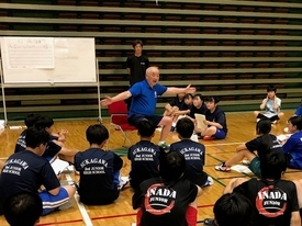 体育館で青いシャツを着た指導者が椅子に座り身振り手振りを交えて熱心に話をしており、背中に学校名のロゴが入った黒いTシャツ姿の学生たちが車座になって聞いている写真