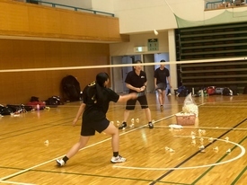 体育館で金色の背番号入り黒Tシャツの参加者が、ネット越しにカゴから次々とシャトルを出す指導者のノック練習を受けており、床に多数のシャトルが散乱している練習風景の写真