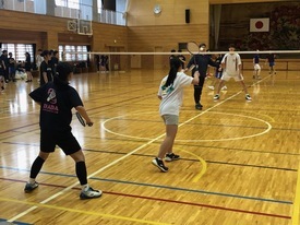 体育館のコート内で手前の黒と白のシャツを着た女子生徒ペアがネット越しに奥の男性ペアと対峙し、バドミントンのダブルスの試合形式で真剣に練習を行っている様子を撮影した写真