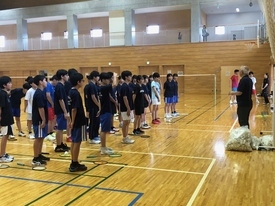 体育館内でスポーツウェアを着た多数の参加者たちが整列し、前に立っている指導者の説明を真剣な表情で聞いている様子を撮影した写真