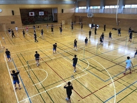 明るい体育館の中で、大勢の参加者たちが、等間隔に張られたいくつものネットを挟んでバドミントンのラケットを構えたりシャトルを打ったりして練習している様子を2階から撮影した写真