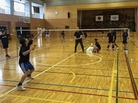 コートで、黒いTシャツと青い短パン姿の生徒がラケットを高く振り上げてシャトルを打つ構えをして、ネットの向こう側では指導者や他の部員たちがその様子を見守りながら練習を行っている写真