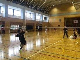 黒い練習着の生徒が躍動感あるフォームでバドミントンのシャトルを打ち、ネット越しに指導者が立っている写真