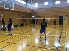 体育館のバドミントンコートで、4人の参加者がネットを挟んでラケットを構え、試合形式の練習を行っている写真