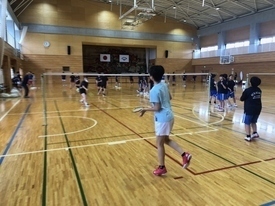 体育館のコートで、水色のTシャツに白い短パンと赤いシューズを着用した男性と参加者の生徒一人が練習を行っている写真