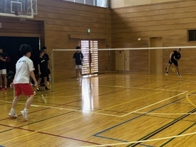 ネットを挟み、手前の白いシャツの男性と奥の黒いシャツの生徒がバドミントンのラリーをしており、コート脇で数名の参加者がその様子を見学している写真