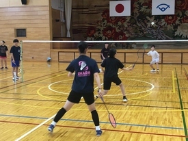 体育館のバドミントンコートで、ペアがネット越しの相手とダブルスの試合をしている様子の写真