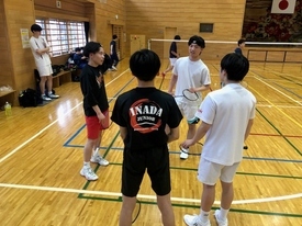 体育館のバドミントンコート上でラケットを持った4人が円になって集まり、指導を行っている様子の写真