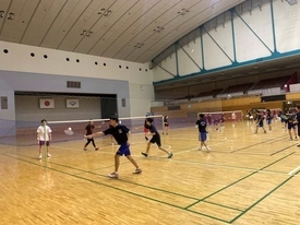 体育館の中に複数のコートが設営され、多くの参加者たちがラケットを構えてシャトルを打ち合いながらバドミントンの練習を行っている様子を撮影した写真