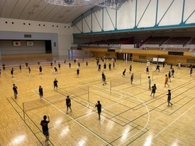 ステージや観客席を備えた広々とした明るい体育館を高い位置から見下ろし、複数のネットが張られたコートで黒いウェアを着た多くの参加者たちが一斉にバドミントンの練習に取り組んでいる様子を2階から撮影した写真