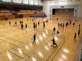 体育館に複数のバドミントンコートが並び、たくさんの参加者たちがラケットを持って練習をしている様子を2階の観客席から撮影した写真