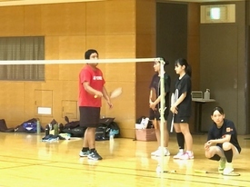 バドミントンコートの横で赤いシャツを着た男性がラケットを構え、その横にラケットを持って立っている女子生徒に指導をしている様子の写真