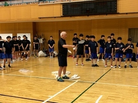 黒いシャツと青いパンツを着た学生たちがラケットを手に整列し、中央にでラケットを持って説明をしている指導者の話を聞いている様子の写真