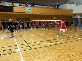 バドミントンコートで赤いシャツを着た男性が整列した参加者の前で実演をしている様子の写真