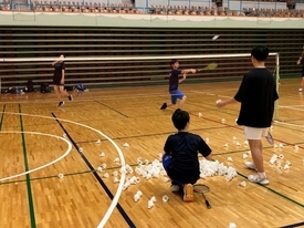 床に沢山のシャトルが置かれているコート側にいる学生が、奥にいる学生に向かってシャトルを打ち込もうとしている写真