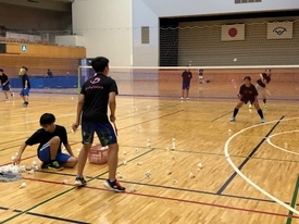 体育館で多数のシャトルが散らばる中、複数の参加者がバドミントンのノック練習に取り組む様子を捉えた写真