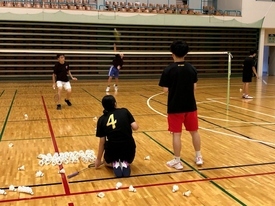 シャトルが散らばるバドミントンコートで、2人の参加者が、ネット越しにノックを受けている様子の写真