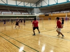 複数のバドミントンコートが並んでいる広い体育館で、赤や黒のシャツを着た参加者たちがネット越しにバドミントンの練習を行っている写真