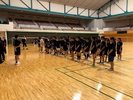 体育館の木製フロアで黒いウェアを着た多数の参加者がラケットを床に置き整列し、指導者の前で説明を受けている様子の写真