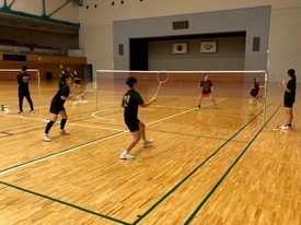 体育館のバドミントンコートで黒いシャツを着た参加者たちがネット越しにラケットを構え、試合形式の練習をしている様子の写真