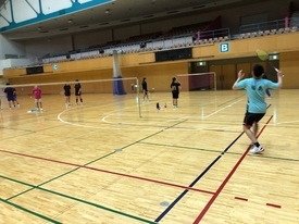 バドミントンコートで、水色のシャツを着た男性がラケットを振り上げ、シャトルを打とうとしている様子の写真