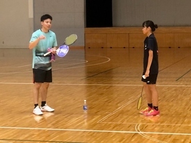 体育館の木製フロアで、水色のシャツを着てラケットを持った男性が、黒いシャツを着た参加者の女子生徒に指導をしている様子の写真