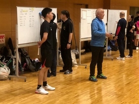 体育館の壁際に置かれた複数のホワイトボードに数人が記入をしており、その前に立った青色の上着の男性が前を向いて話をしている写真