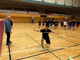 体育館で黒いウェアを着た男子生徒がラケットを振り、前に立った青色の上着の男性が指導をしている様子の写真