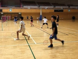 体育館に複数のバドミントンコートが並び、黒や白のウェアを着た参加者たちがラケットを持って練習をしている様子の写真