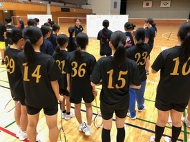 背番号入りの黒いシャツを着た参加者たちが、ネットの前に置かれたホワイトボード前に集まっている写真