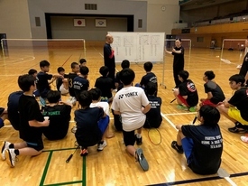 コートの中に置かれたホワイトボード前に集まった参加者たちが、座って指導者の説明を聞いている写真