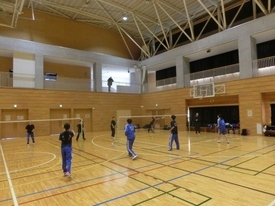 ネットが張られた2面のコートとその脇で、複数の参加者がバドミントンの練習をしている様子の写真