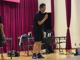 紫色のカーテンが引かれたステージ上で、黒いポロシャツとハーフパンツ姿の男性がマイクを手に持ち、話をしている写真