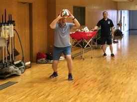 ポロシャツを着た男性がバレーボールを頭上に構えてトスのフォームを確認しており、背後で紺色のシャツを着た男性がその様子を真剣な表情で見守っている練習風景の写真