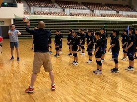 黒いユニフォームを着た大勢の生徒たちが整列している前で、2人の男性がボールの扱いの見本を見せている様子の写真