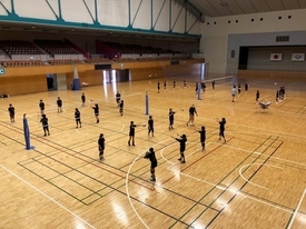 広い体育館のコート内で、黒いウェアを着用した大勢の生徒たちが等間隔に並び、一斉にバレーボールの基礎フォームを練習している写真