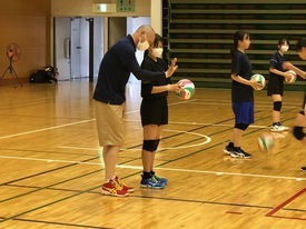 体育館のコート内で、マスクを着用した指導者の男性が女子生徒の隣に立ち、フォームを丁寧に指導している写真