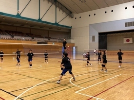 体育館のコート内で、黒いウェアを着用した大勢の生徒たちがネットを挟んで複数のグループに分かれて練習を行っている写真