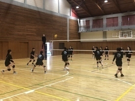 体育館のコート内で、黒い練習着の生徒たちが試合形式のバレーボールの練習をしている写真