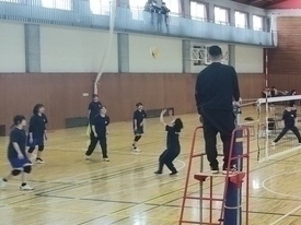 体育館のコート内で、生徒たちがバレーボールをトスを行っており、その様子を黒いウェアを着た指導者が見守っている写真