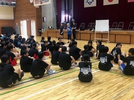 木製フローリングの体育館で、多数の生徒たちが黒い服装で正座し、ステージ前で指導者の方々が説明している様子の写真