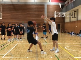 体育館の中で、一人の生徒がドリブルをしながらもう一人の生徒にバスケットボールをパスしようとしている瞬間を捉え、周りには多くの生徒がボールを持って練習を見守っている写真