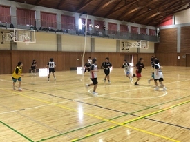 体育館の中で、木目の床と壁を背景に、生徒たちがユニフォーム姿でバスケットボールの試合を行っており、コート上でボールを追いかけ走っている様子の写真