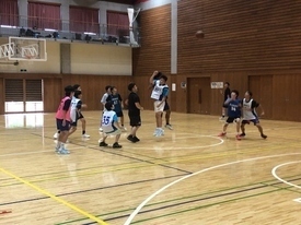 体育館の木目のフロアで、複数の生徒がユニフォームや練習着を着用し、一人の生徒がジャンプしてシュートを打とうとし、他の生徒たちがディフェンスやリバウンドの位置についている写真