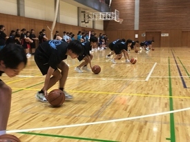 体育館の木製フロアで、多数の子どもたちが黒いユニフォームを着てバスケットボールのドリブル練習をしている写真