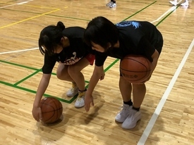 体育館の木製フロアで、黒いトップスを着た2人の子どもが腰をかがめてバスケットボールを使ったドリル練習をしている様子の写真