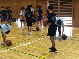 体育館の木製フロアで、紺色のユニフォームを着た子どもたちがバスケットボールのドリブル練習をし、背景に他の参加者が見ている様子の写真