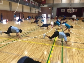 体育館の木製フロアで、青や黒のスポーツウェアを着た子どもたちが列になって腕立て伏せなどの体幹トレーニングをしている写真