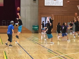 体育館の木製コートで、様々な色のスポーツウェアを着た子どもたちがボールを上に投げゴール練習をしている写真