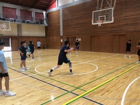 体育館の木製コートで、紺や黒のスポーツウェアを着た子どもたちがバスケットゴールに向かってシュート練習をしている様子の写真